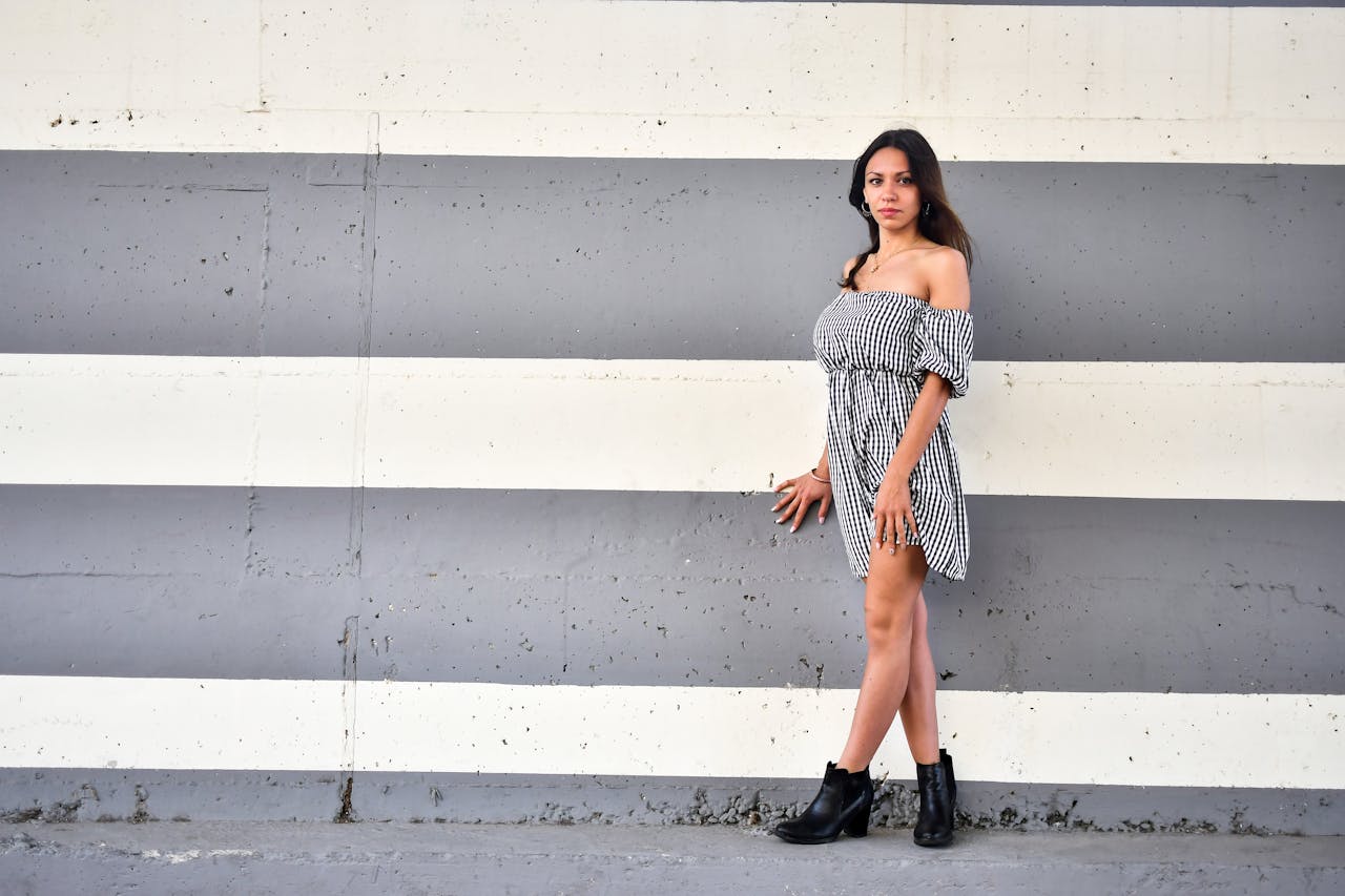 A fashionable woman in an off-shoulder dress poses stylishly against a striped wall.